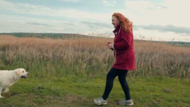 Kızıl saçlı bir kadın, güneşli bir günde, uzun otlarla ve bulutlu gökyüzüyle çevrili çimenli bir alanda iki köpeğiyle, siyah bir laboratuvarla ve beyaz bir laboratuvarla oynar..