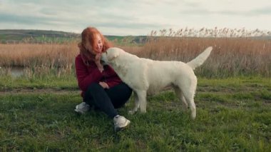 Neşeli bir kadın sakin bir gölün yanında neşeli köpeğiyle çimlerin üzerinde oturur. Açık havada güzel bir günün tadını çıkarırlar yemyeşil ve hafif meltemler arasında..