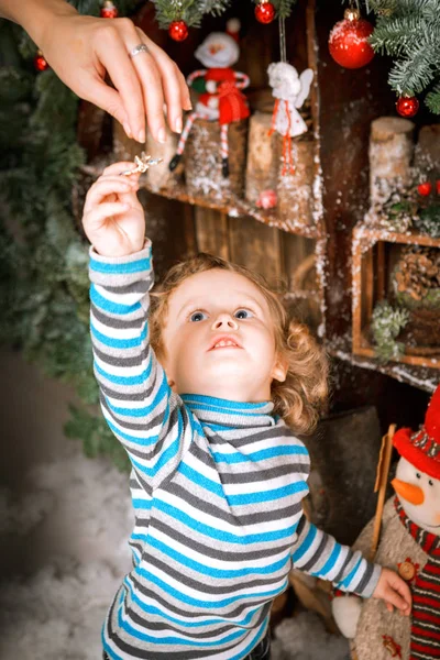 Niño de dos años vestido con chaqueta de cuero braun, pantalones y botas con sombrero piloto navidad árbol de Navidad en decoraciones de madera — Foto de Stock