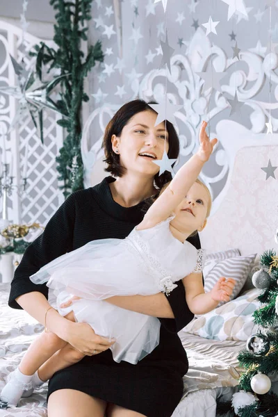 Feliz madre sostiene hija niña en sus vueltas dando un regalo y blanco conejito cerca de árbol de Navidad en blanco clásico interior — Foto de Stock