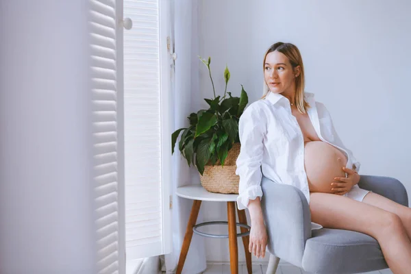 怀孕的 Mothercare 生活方式的概念 鲍勃发型金发女郎的健康护理 白色的灯光内饰 母亲坐在灰色的扶手椅上 房间里带着眼罩和木地板 绿色植物在角落里 — 图库照片