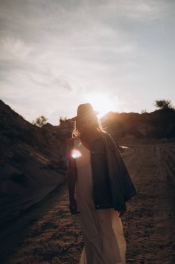 Şapkalı, beyaz elbiseli, kot ceketli güzel sarışın kadın çölde yürüyor.