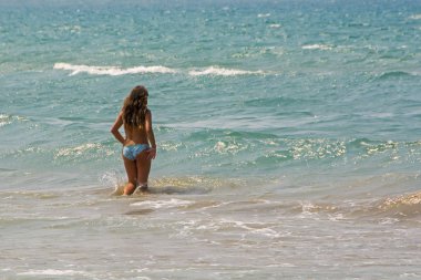 Masmavi deniz parlak yaz gün mavi bikini duran dalgalar ile güzel genç kadın. Seksi kız deniz sahilinde arkasında. Kumlu sahilde rahatlatıcı tabaklanmış kadınla