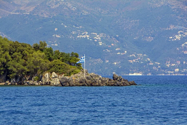 ギリシャ国旗 ひびの入った岩崖のマウントに緑の熱帯の木々 とコルフ ギリシャの美しい小島 ターコイズ ブルーの背景に青い山と海の — ストック写真