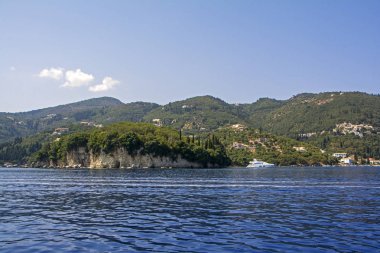 Masmavi denizi Corfu Island, Yunanistan koyu yeşil ağaçlar ve villalar arka plan üzerinde beyaz yat demirleyen