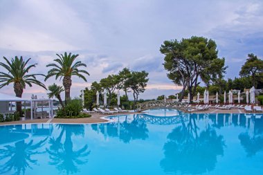 Yeşil ağaçlar, şemsiye, şezlong ve yüzme havuzu ve güzel gün batımını karşı sakin su yansıması üzerinde ahşap vintage köprü ile sakin sahne