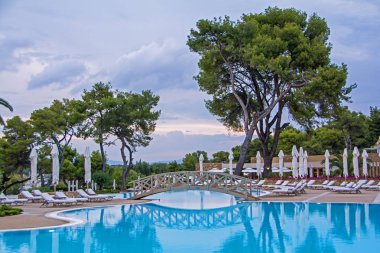 Yeşil ağaçlar, şemsiye, şezlong ve yüzme havuzu ve güzel gün batımını karşı sakin su yansıması üzerinde ahşap vintage köprü ile sakin sahne