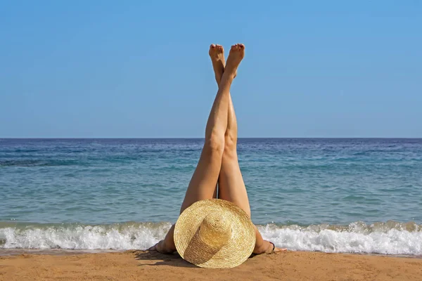 Güzel genç kadın ince bacaklar uzanan tropikal bir kumsalda yatıp hasır şapkalı. Arka plan bulanık mavi denizde. Yaz tatil kavramı