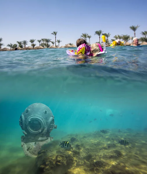 Vintage antique deep sea diving metal helmet on sea bottom near reef with exotic fishes and the kid in diving mask examines underwater world. Beautiful beach with palm trees and blue sky on background