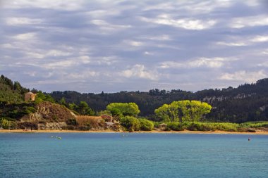 Erikousa adasının güzel manzarası, geleneksel eski yel değirmeni ile Yunanistan, deniz mavisi, Güneş ve mavi bulutlu gökyüzü ile aydınlatılmış yeşil ağaçlar