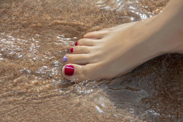 Nice woman leg with red pedicure on the wet sand in a sea water on sandy beach