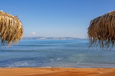 Saman plaj şemsiyeleri, kum sahili ve berrak gökyüzünün altında mavi deniz ile güzel bir sahne. Doğal çerçeve, tasarım ve metin için mükemmel arka plan