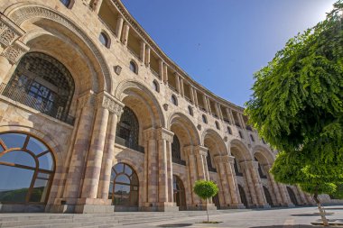 Cumhuriyet Meydanı, Erivan, Ermenistan'da ön planda yeşil ağaçlarla mavi gökyüzüne karşı kemer ve sütunlarla tüften yapılmış güzel klasik binaların düşük geniş açılı görünümü