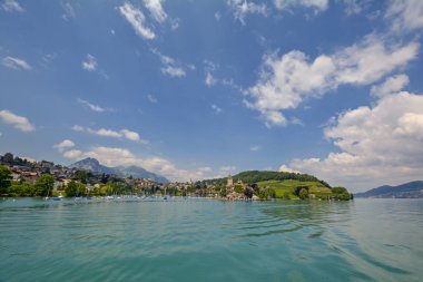 İsviçre'de mavi bulutlu gökyüzü altında demirlemiş lüks yatlar, yeşil tepeler ve İsviçre Alpleri ile Thun Gölü kıyısında antik Spiez Kalesi (Schloss Spiez) ile güzel manzara