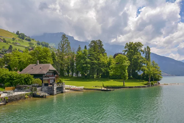 スイスの青い曇りの空の下に緑の丘とスイスアルプスとトゥーン湖の海岸にタイル屋根と伝統的な木製の建物と美しいカラフルな風景 — ストック写真