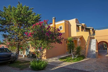 Yunanistan 'ın Corfu adasındaki deniz kıyısında, mavi gökyüzünün altında lüks kulübeleri, yeşil ağaçları ve pembe çiçekleri olan güzel bir bahçe.
