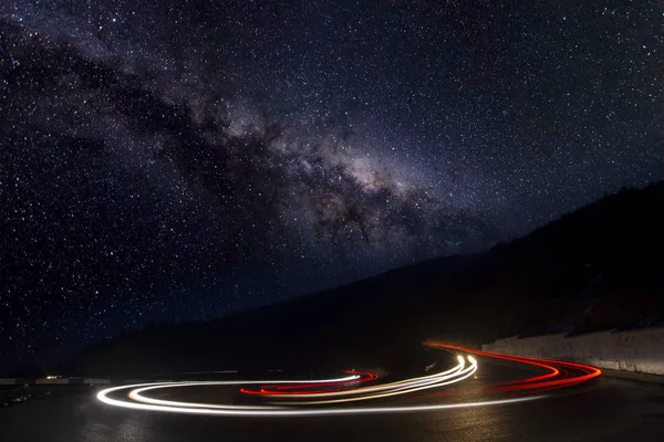 Milky Way Galaxy over mountain road at night. Blurred car headlights on winding road. Colorful landscape with sky with stars and blue milky way