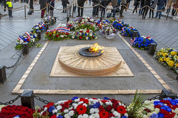 Paris, France - November 17, 2018: Eternal flame of the Tomb of the Unknown Soldier surrounded with flowers and wreaths in colors of French flag under the Triumphal arch (Arc de Triomphe du Carrousel)