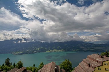 Thun Gölü 'nün panoramik manzarası (Thunersee) turkuaz su ve İsviçre Alpleri ile Beatenberg, İsviçre' den dramatik bulutlu gökyüzü altında binaların kiremit çatıları ve ön planda yeşil ağaçlarla