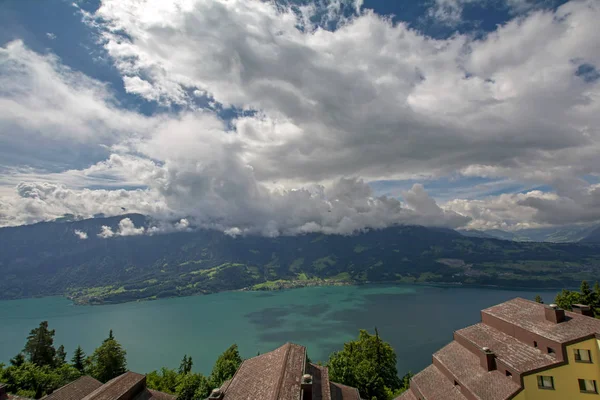 ビーテンバーグからのターコイズブルーの水とスイスアルプスとのトゥン湖 Thunersee のパノラマビュー 建物のタイル屋根と前景に緑の木と劇的な曇り空の下でスイス — ストック写真