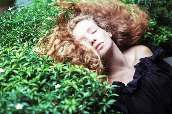 Beautiful young red curly hair woman relaxing on the bushes in the park