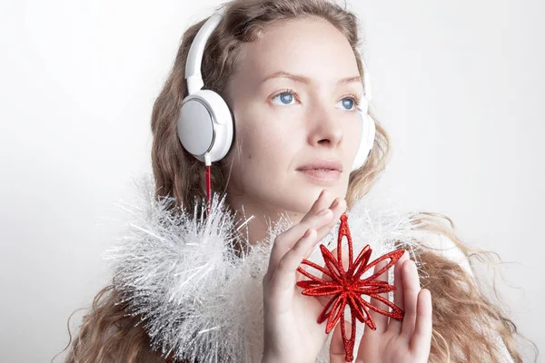 smiling redhead woman in white scarf listening music with headphones in front of white wall and holding red decorative snowflake, christmas concept