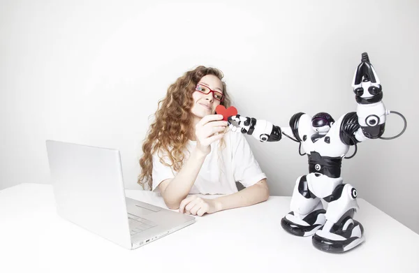 微笑的红头发的妇女与笔记本电脑在白色的墙壁前工作 而机器人给她的红心 — 图库照片