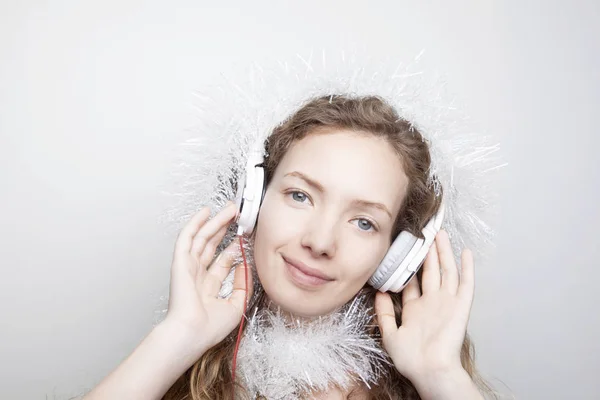 smiling redhead woman in white scarf listening music with headphones in front of white wall and looking at camera