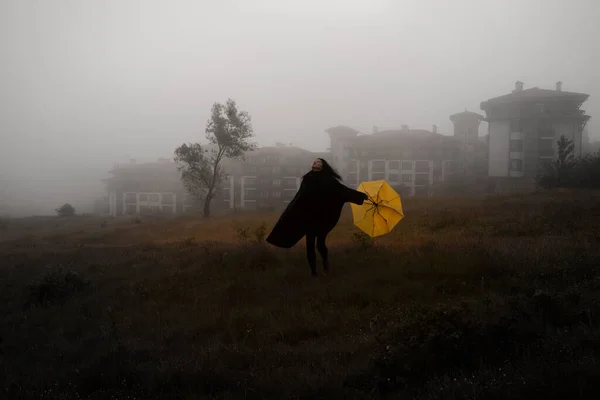 穿着黑色夹克的寂寞女人带着黄色的雨伞在森林的雾中走着 — 图库照片