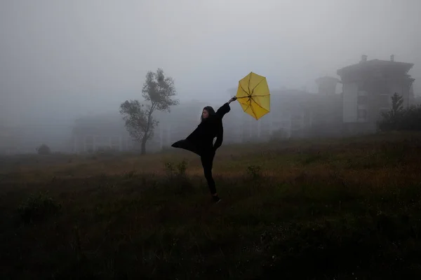 穿着黑色夹克的寂寞女人带着黄色的雨伞在森林的雾中走着 — 图库照片