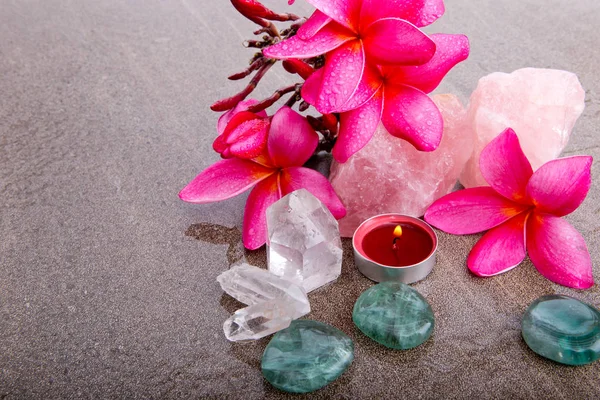 Pink Frangipani flowers with healing crystals (rose quartz, clear quartz, flourite) and red candle on wet brown slate background