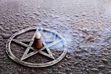 Burning incense, with wisps of smoke, in center of gray metal pentagram on slate background with water drops - with copy space clipart