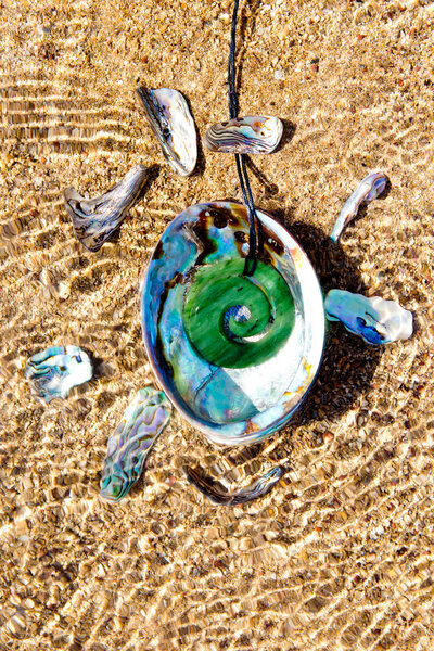 Новая Зеландия - Маори тема - Джейд pounamu Koru дизайн кулон с Paua скорлупа лежит на мелководье на пляже - абстрактно с рябью воды и солнечного света
