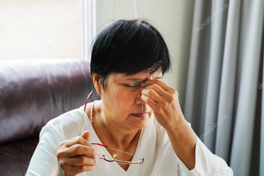 Tired old woman removing eyeglasses, massaging eyes after reading paper book. feeling discomfort because of long wearing glasses, suffering from eyes pain or headache