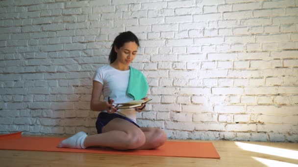 Menina bonita escreve um programa de treinamento de fitness. Contra uma parede de tijolo branco — Vídeo de Stock