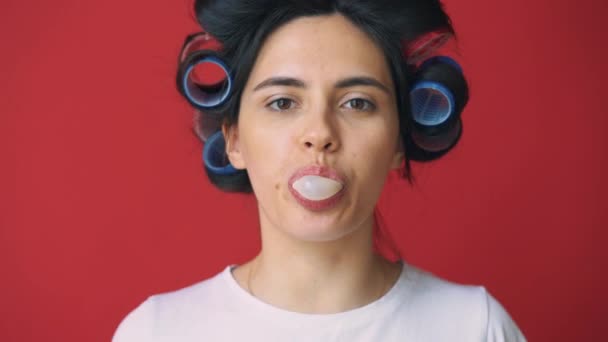 Flicka med papiljotter på hennes huvud och tuggummi blåser bubble på röd bakgrund. — Stockvideo