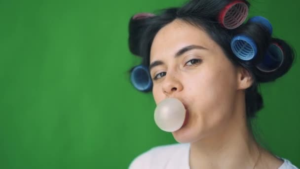 Chica con rulos en la cabeza y goma de mascar, burbujas que soplan sobre un fondo verde, cromakey . — Vídeos de Stock