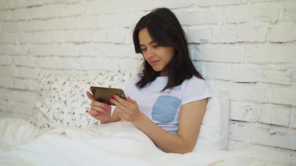 Vue latérale d'une jeune femme couchée au lit et bavardant en vidéo avec son smartphone — Video