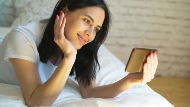 Vista lateral de la mujer joven acostada en la cama y video chat con su teléfono inteligente — Vídeos de Stock