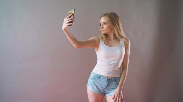 Mooi meisje met lichte make-up neemt een selfie op een grijze achtergrond. — Stockvideo