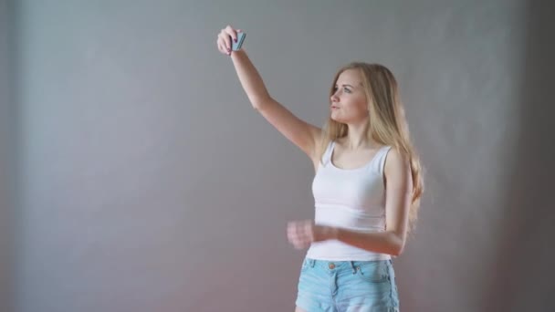 Beautiful girl with light makeup takes a selfie on a gray background. — Stock Video