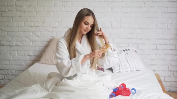 Hermosa chica sentada en el dormitorio en la cama retuerce los rizadores, cabello hermoso y saludable — Vídeos de Stock