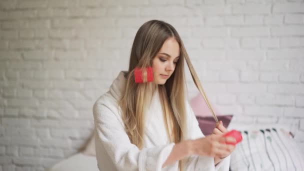 Hermosa chica sentada en el dormitorio en la cama retuerce los rizadores, cabello hermoso y saludable — Vídeo de stock