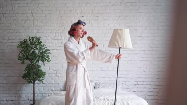 Glückliche junge blonde Frau mit ziemlich langen Haaren im Pyjama springt und tanzt morgens auf dem Bett und lächelt. Frau in fröhlicher Stimmung — Stockvideo