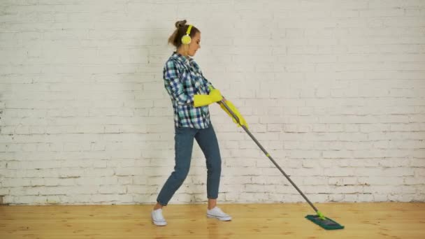 A young woman with headphones and a MOP is ready for home cleaning. Dancing and singing to good music — Stock Video