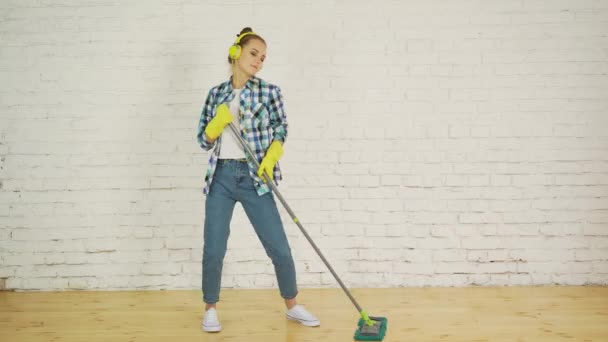 Uma jovem com fones de ouvido e um MOP está pronta para limpeza em casa. Dançando e cantando a boa música — Vídeo de Stock