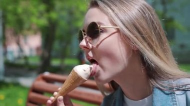 Genç kız elleri, dondurma ile bir bankta oturan beyaz t-shirt, kot, kot pantolon ve güneş gözlüğü giyiyor