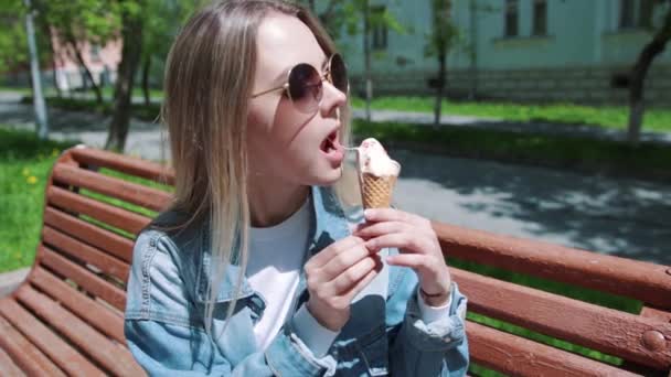 Young girl sitting on a bench with ice cream on her hands, wearing a white t-shirt, denim, jeans and sunglasses — Stock Video