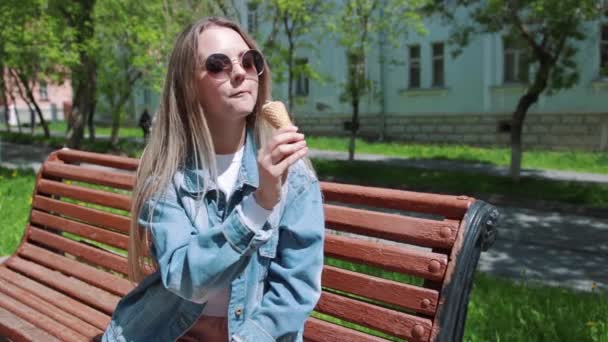 Giovane ragazza seduta su una panchina con gelati sulle mani, indossando una t-shirt bianca, denim, jeans e occhiali da sole — Video Stock