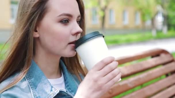 Concept de boissons et de personnes - jeune femme heureuse ou adolescente buvant du café dans une tasse en papier assise sur un banc de rue de la ville — Video